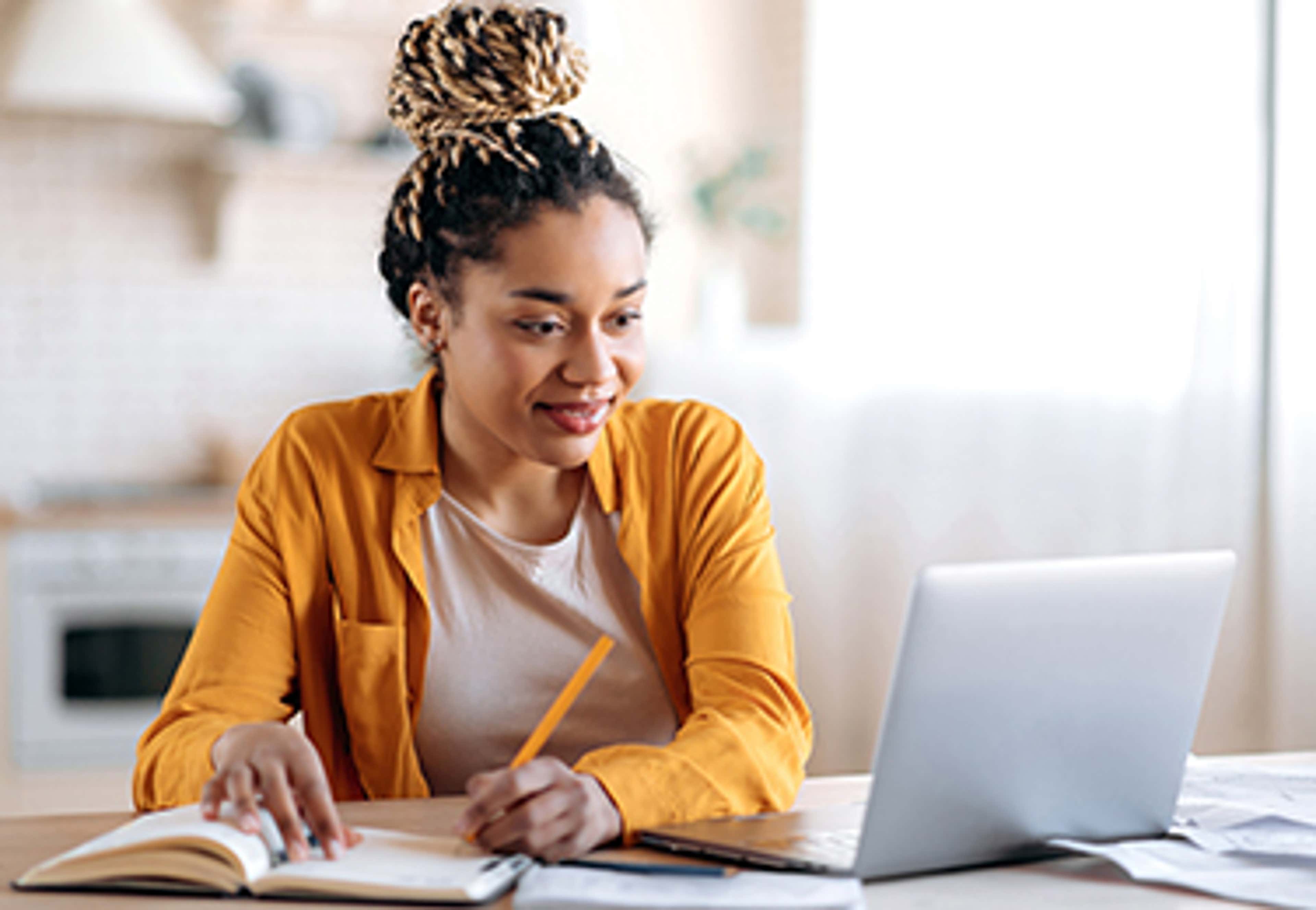 Young person sitting at desk with laptop, writing in a book