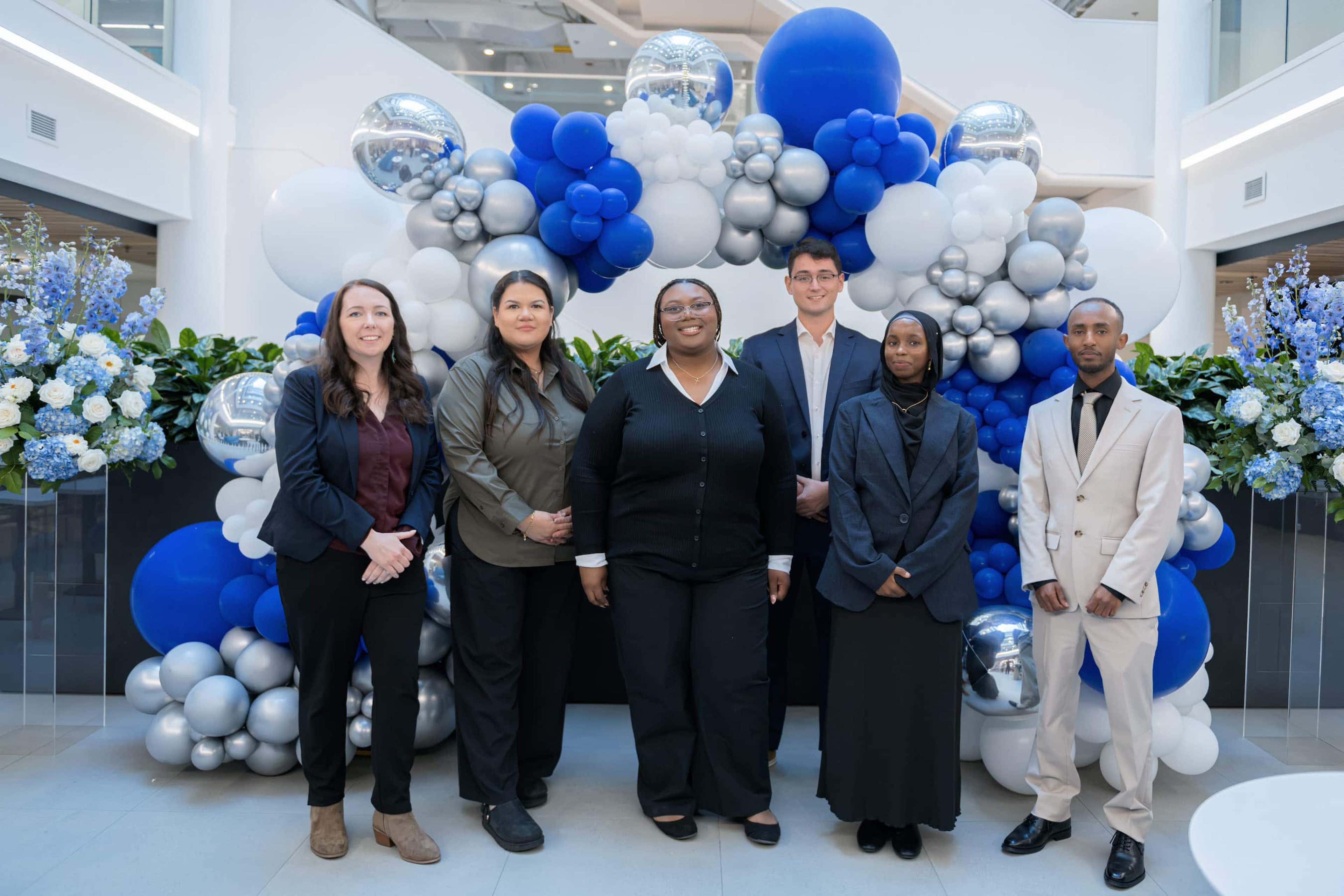 A diverse group of people smiling and standing together in front of a blue and white balloon arch.