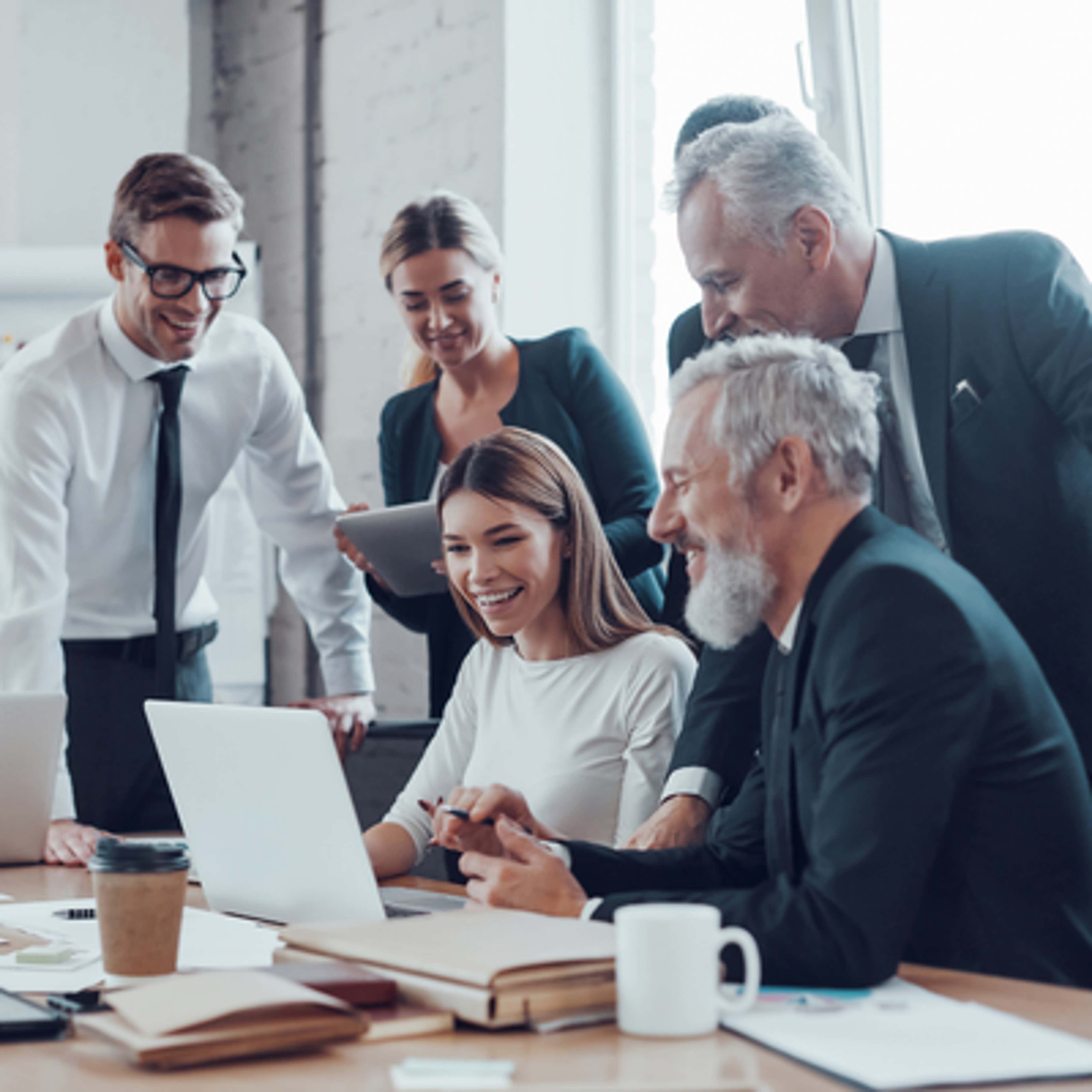 A group of business professionals collaborates around a laptop, discussing ideas and strategies in a modern office setting.