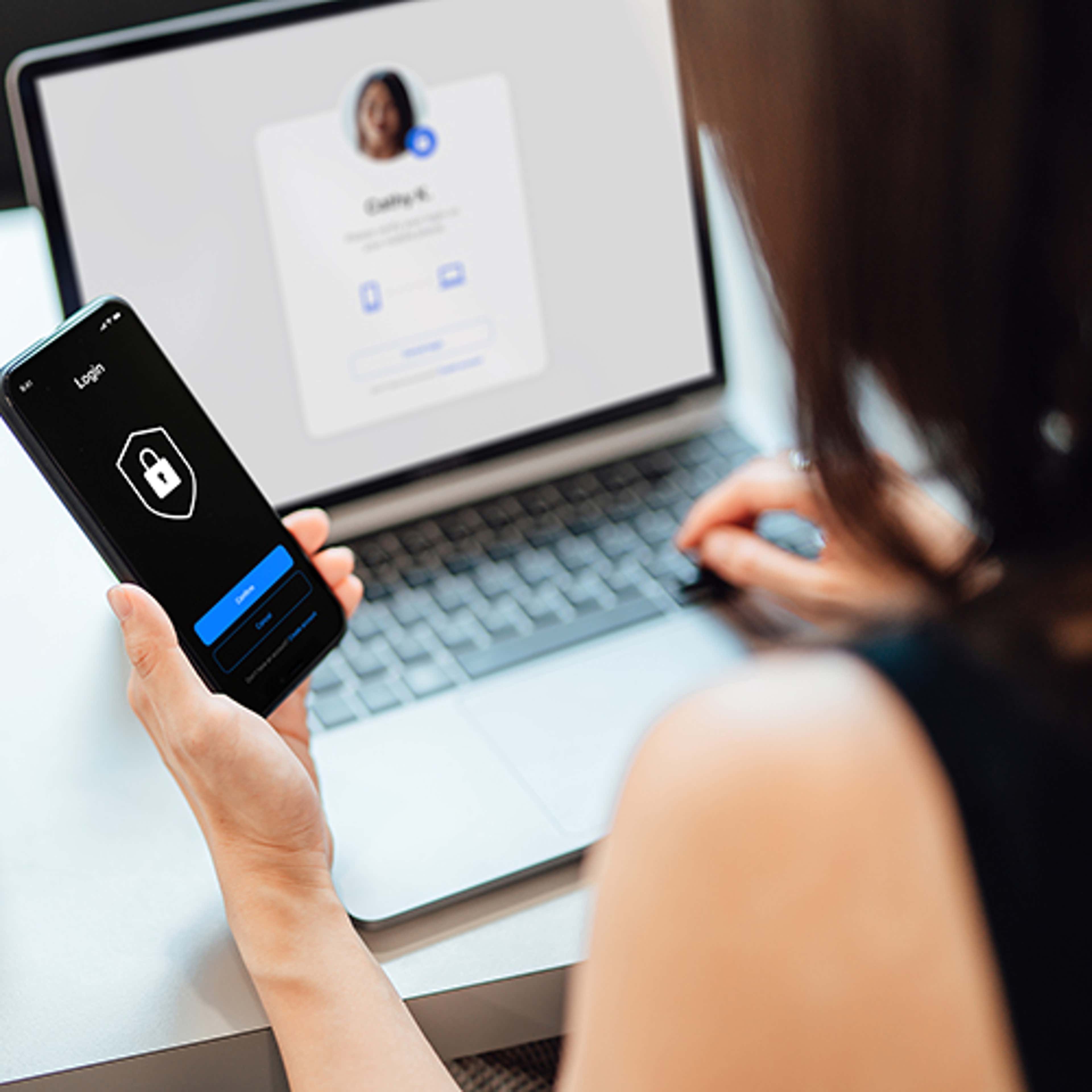 Person sitting at laptop with phone in hand, doing multi-factor authenication