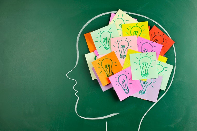 Head drawn in white chalk on a green chalkboard with post-it notes with lightbulbs drawn on them placed within the head.