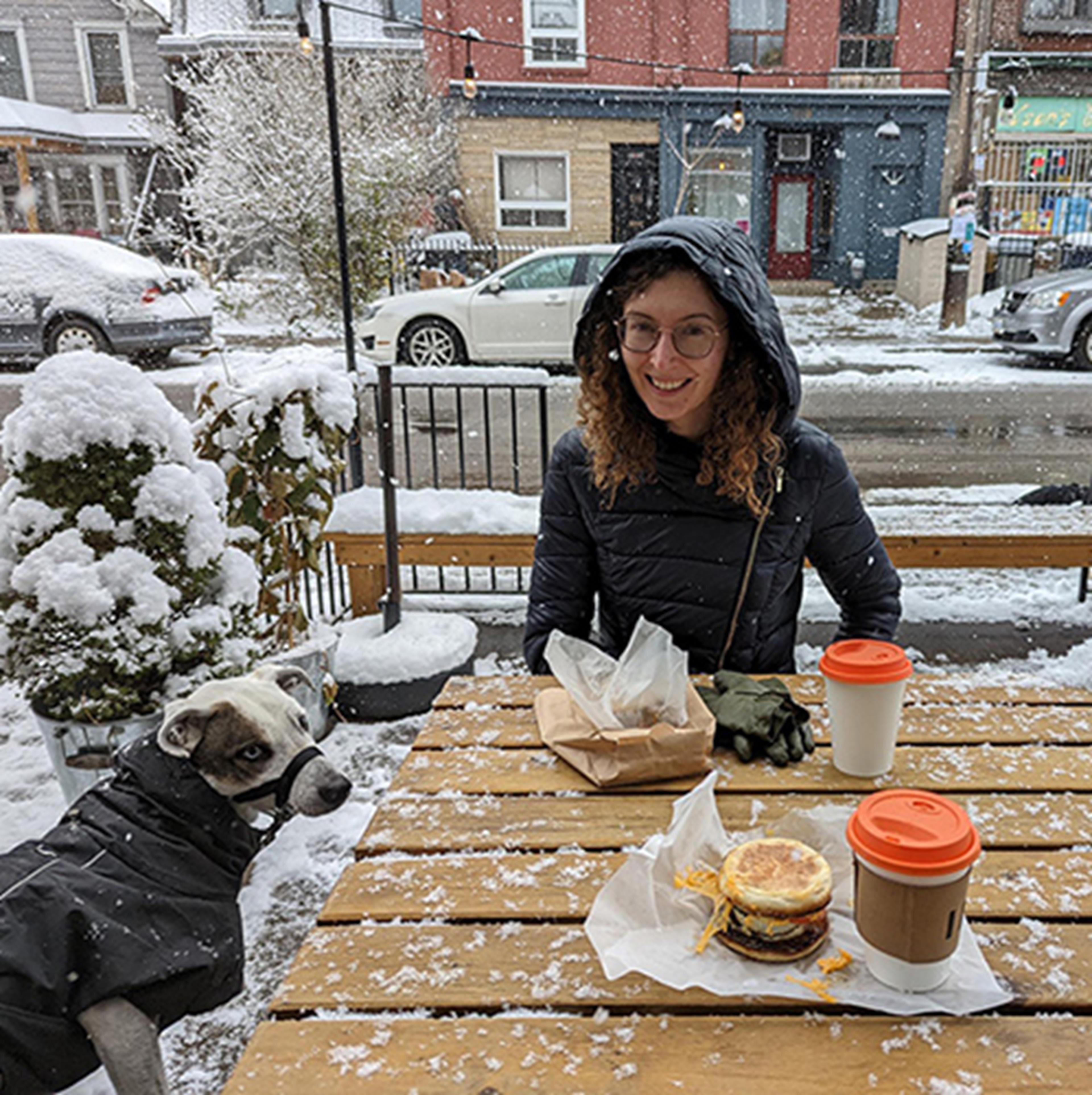 Madison sitting at an outdoor table in the winter with a cup of coffee and a dog
