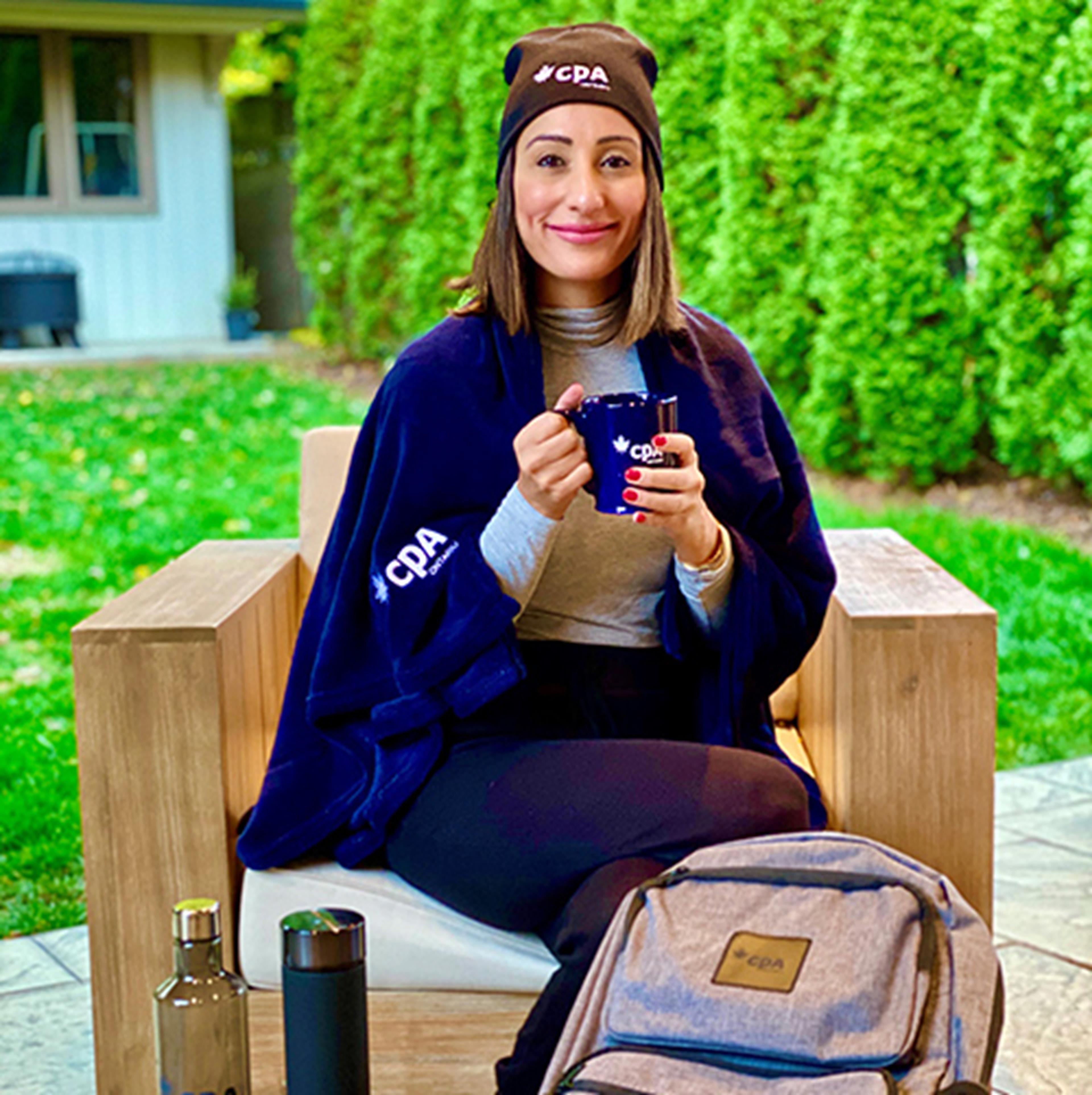 Narmin sitting on a chair outside with a CPA hat, mug and blanket