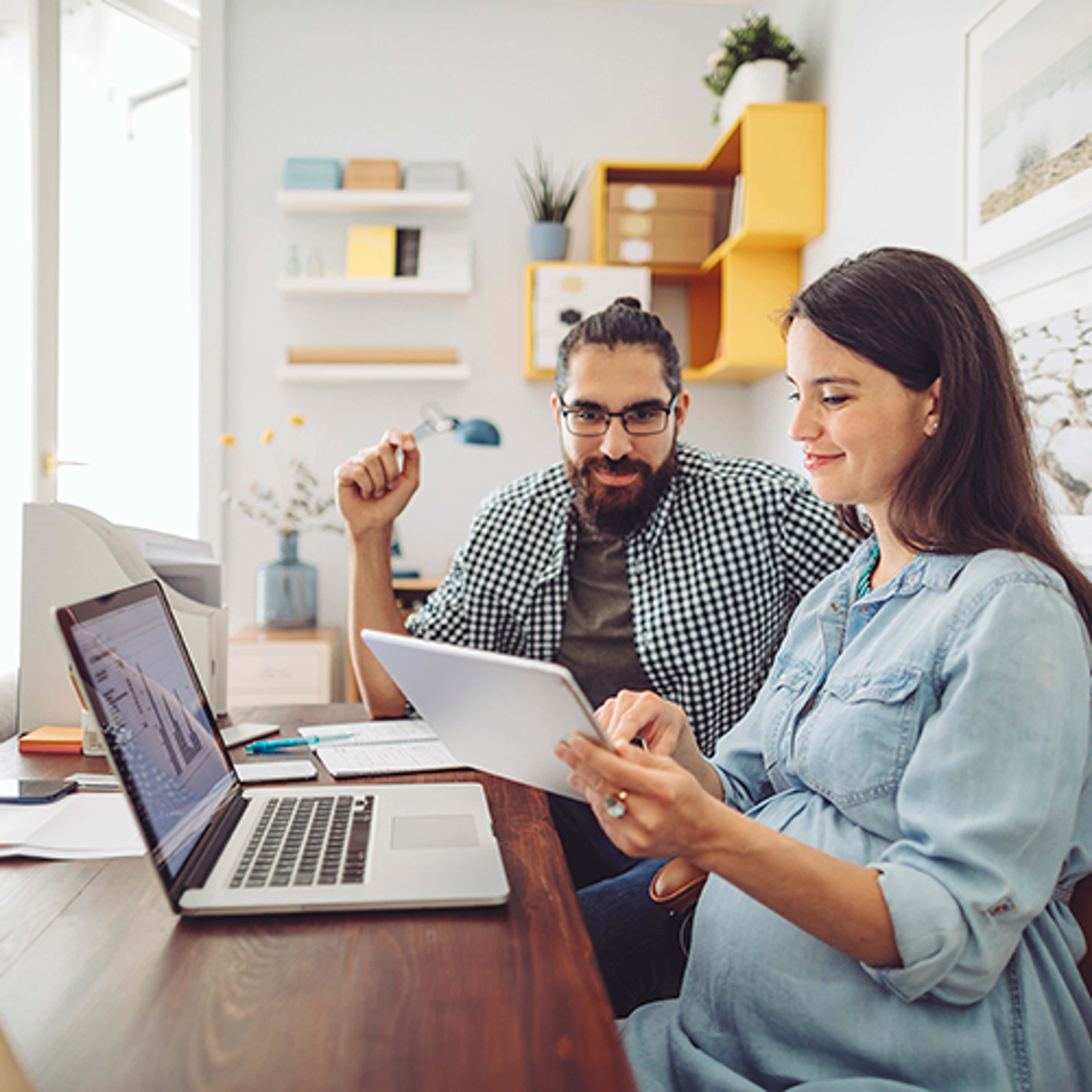 Man with pregnant woman sitting at table looking at tablet and laptop
