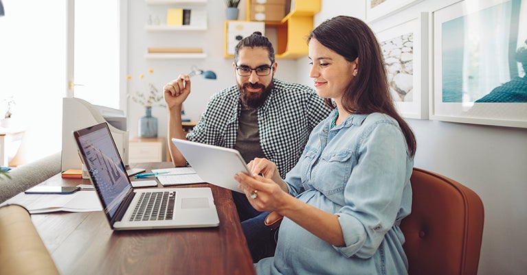 Man and pregnant woman sitting on living room couch looking at tablet and laptop