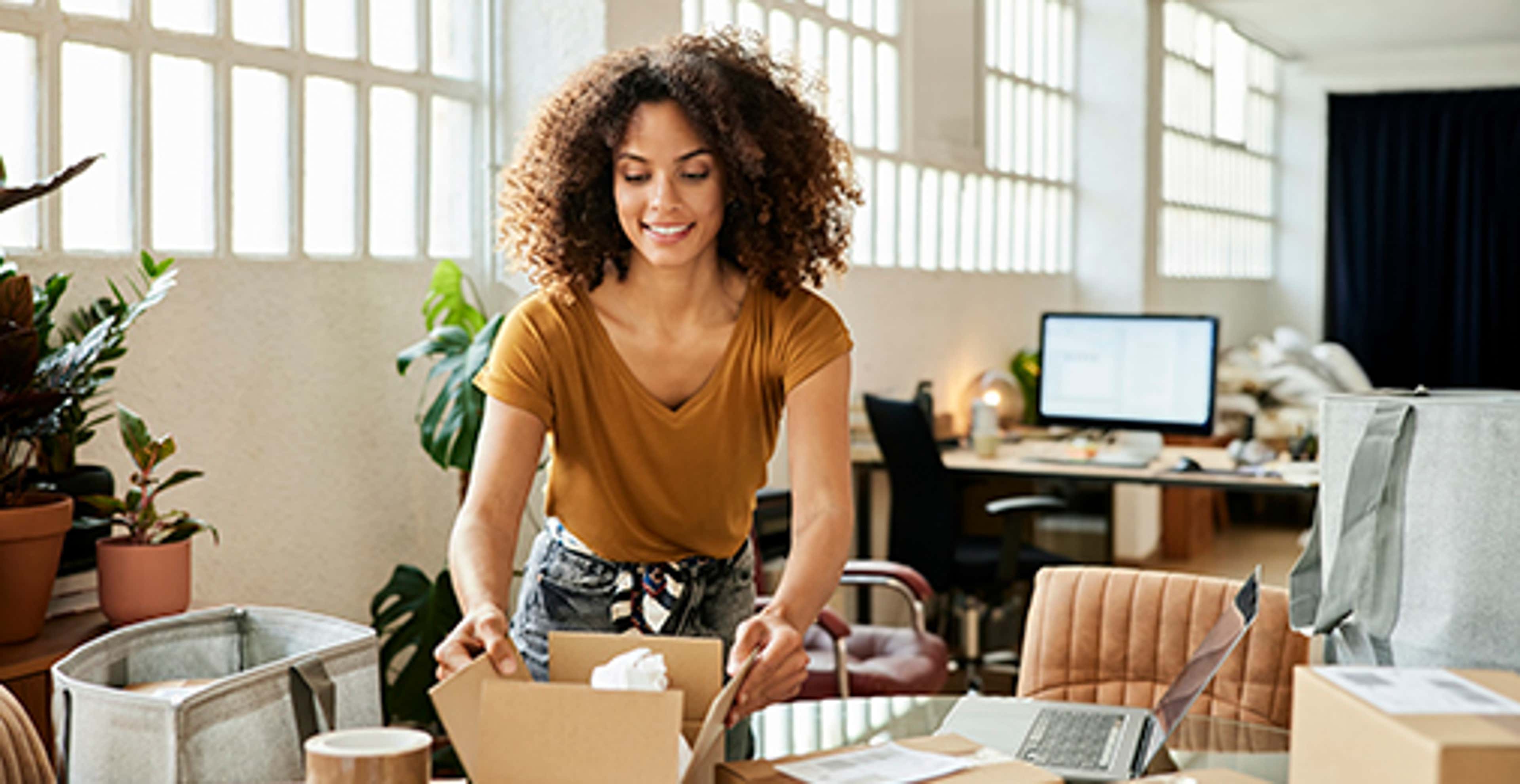 Woman in an open space office standing up over a desk holding a box