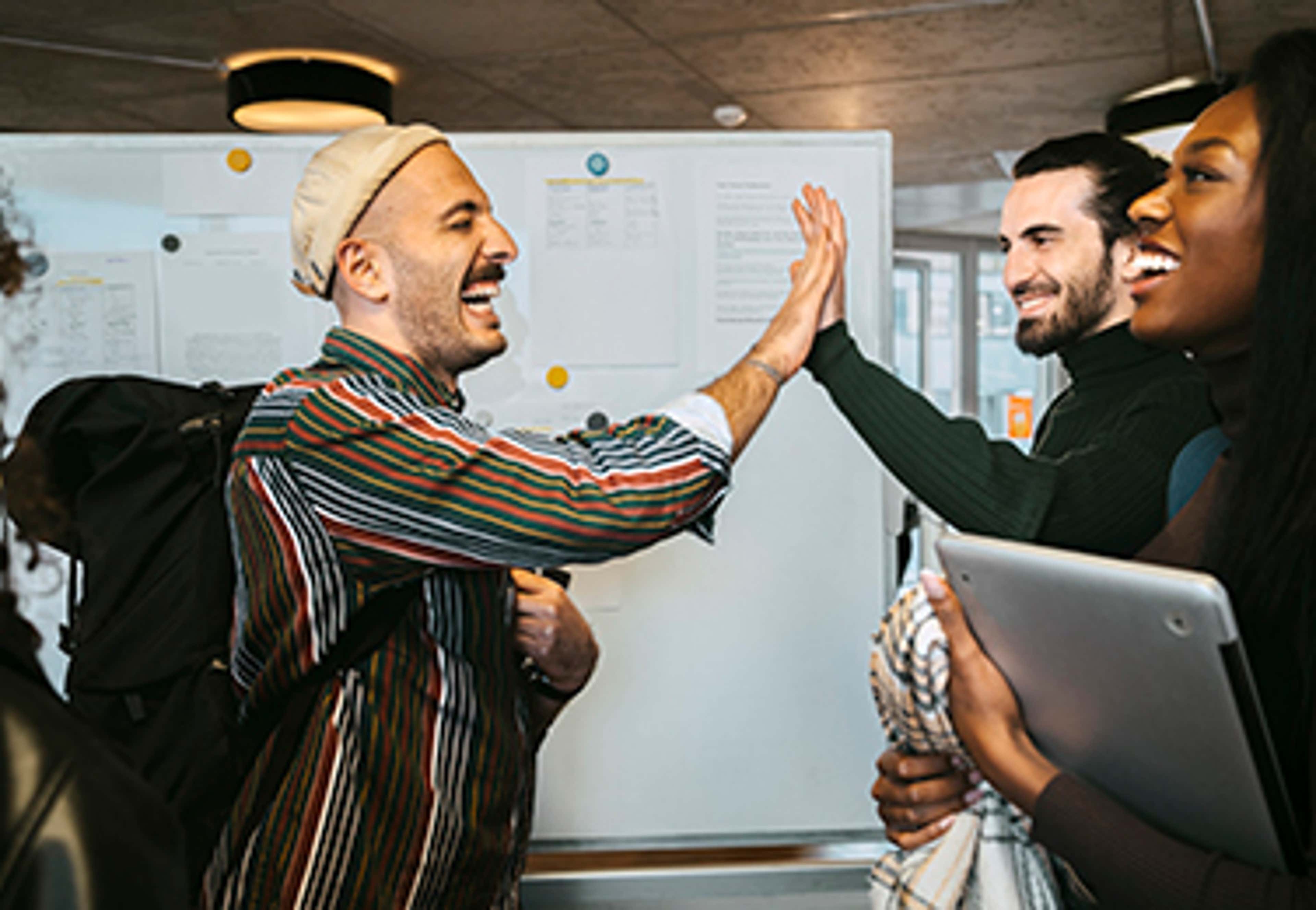 Person with striped shirt, backpack and hat giving a high-five to another person in front of a bulletin board.