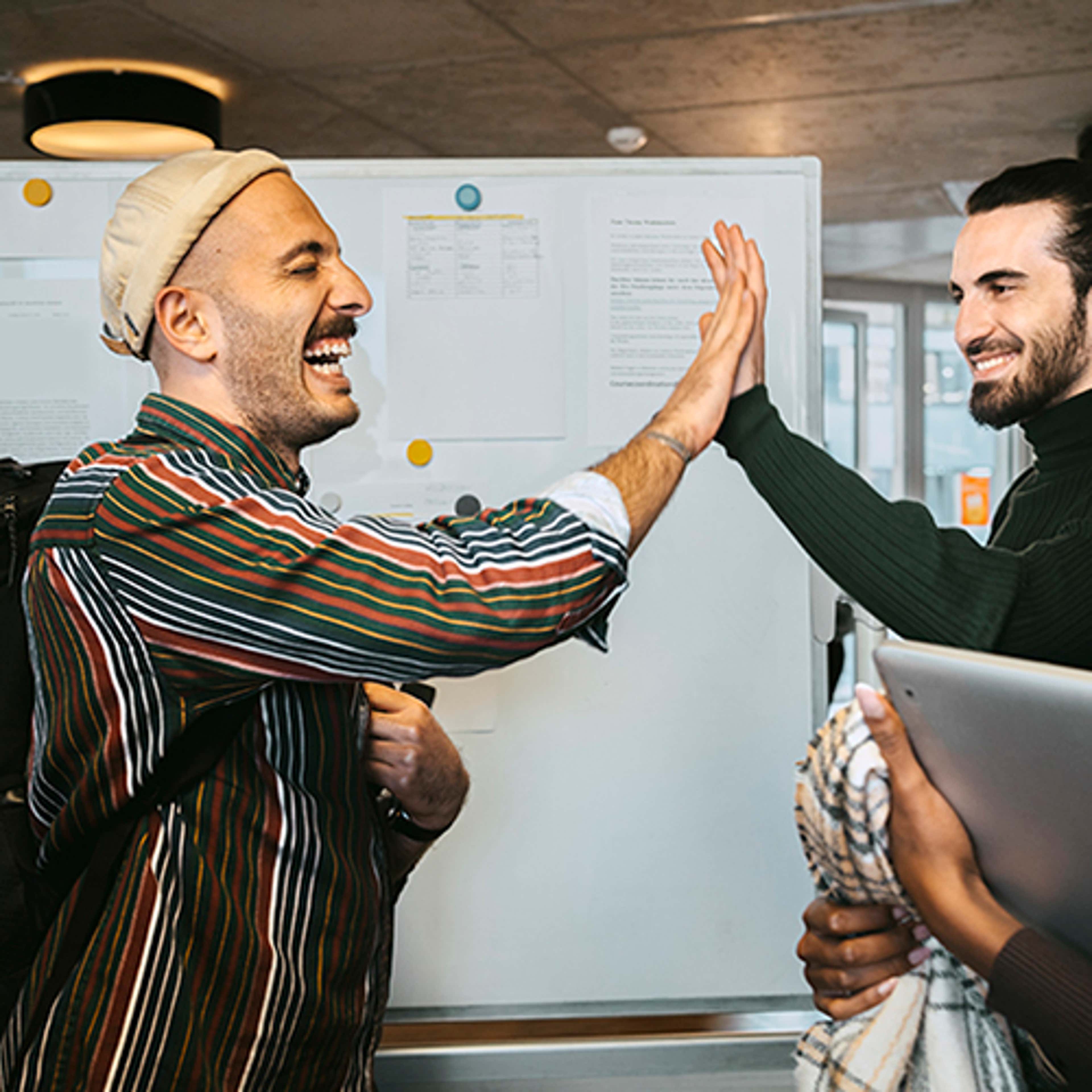 Person with striped shirt, backpack and hat giving a high-five to another person in front of a bulletin board.