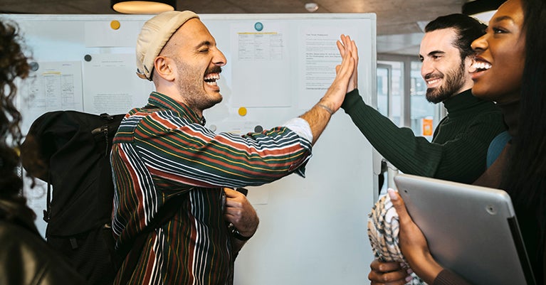 Person with striped shirt, backpack and hat giving a high-five to another person in front of a bulletin board.