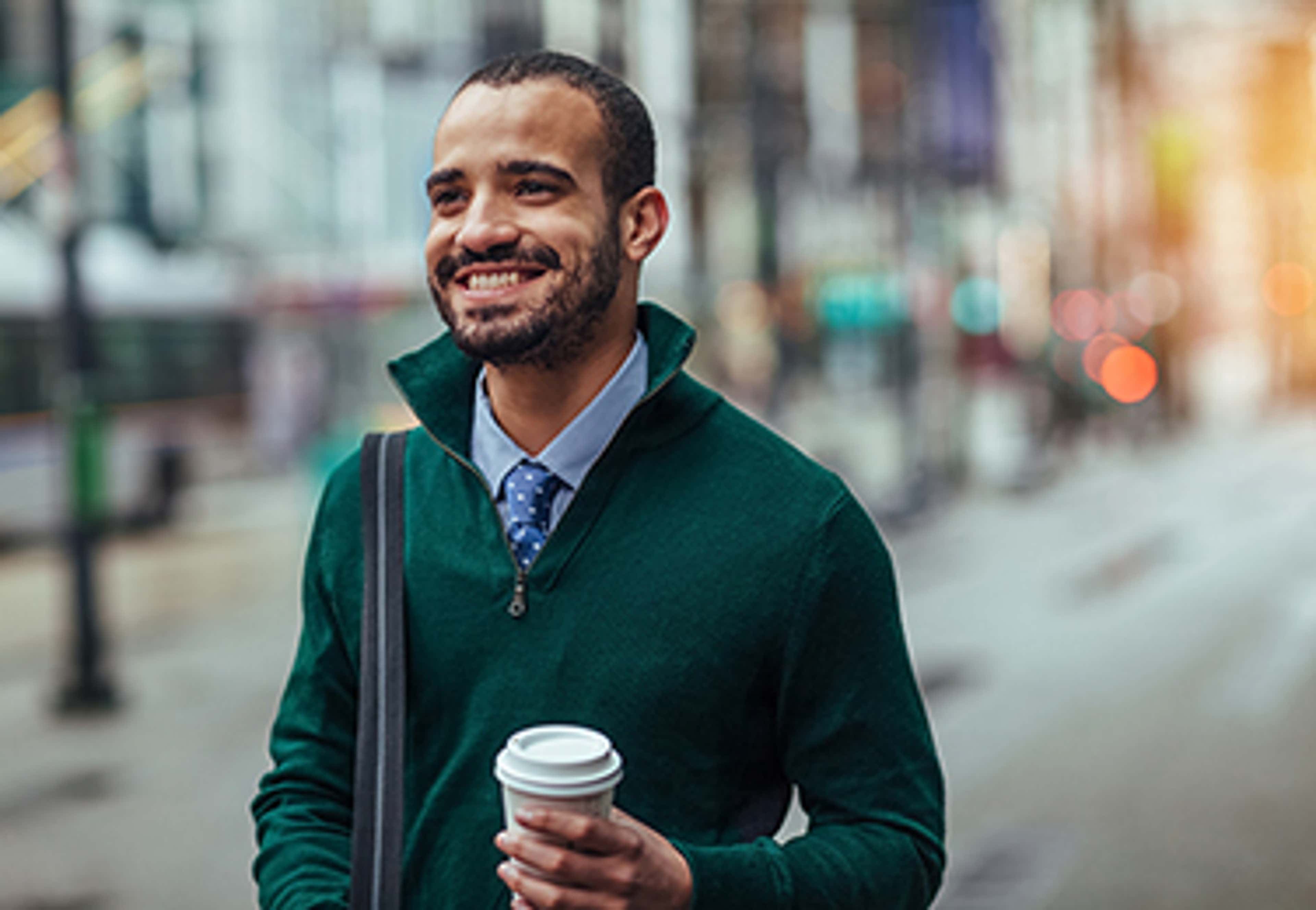 Man with green sweater, walking and smiling with coffee in hand