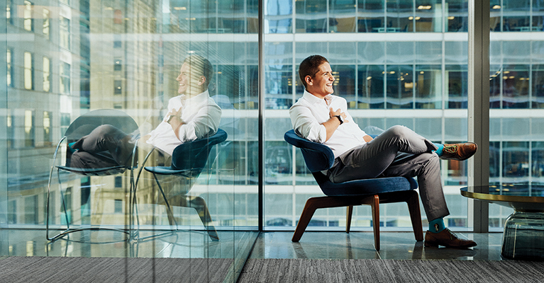 Michael sitting in a chair in front of a floor to ceiling glass window, looking away from the camera and smiling