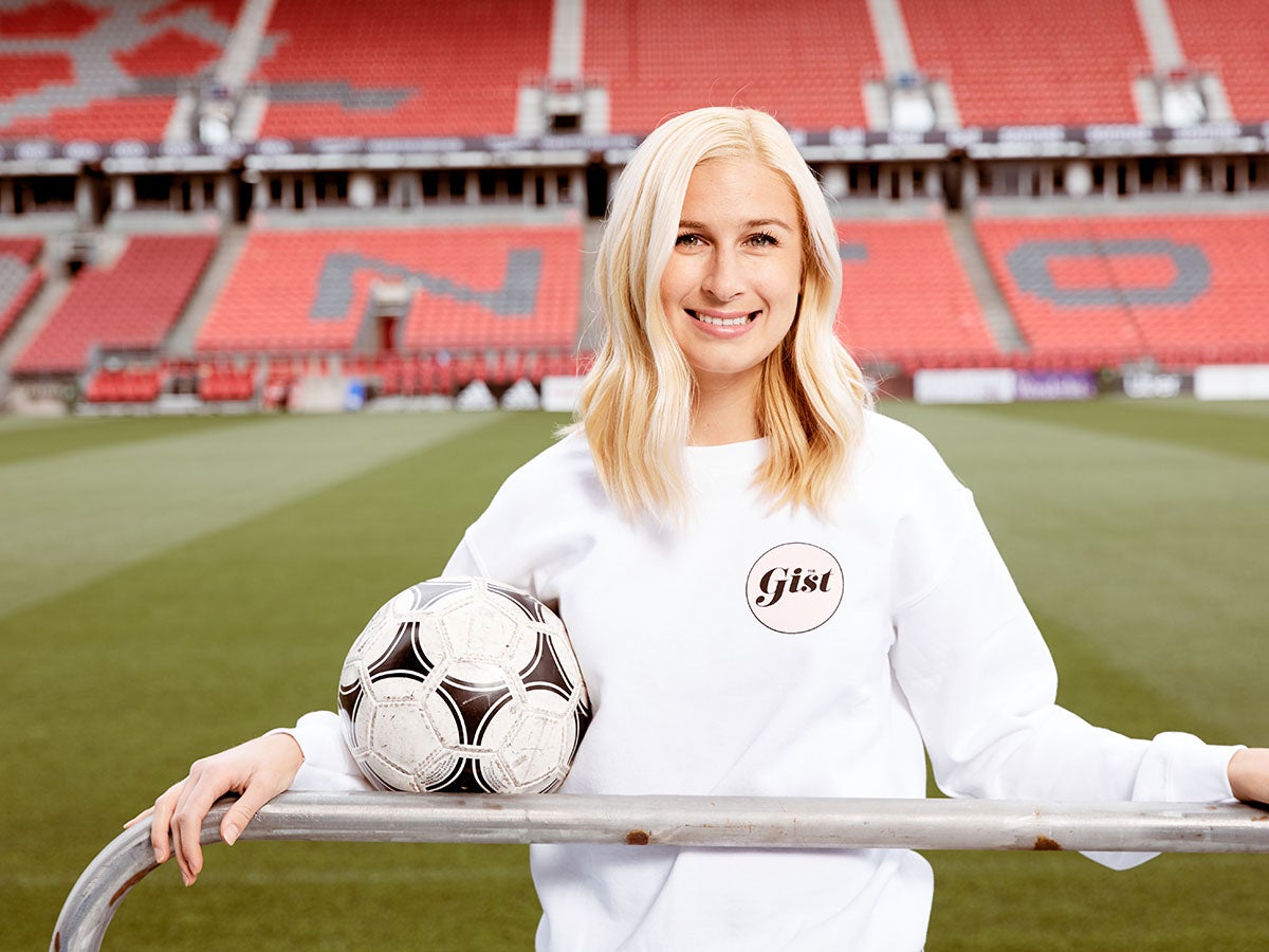 Roslyn Mclarty standing in a stadium, holding a soccer ball and wearing a white jersey with a Gist logo on the left