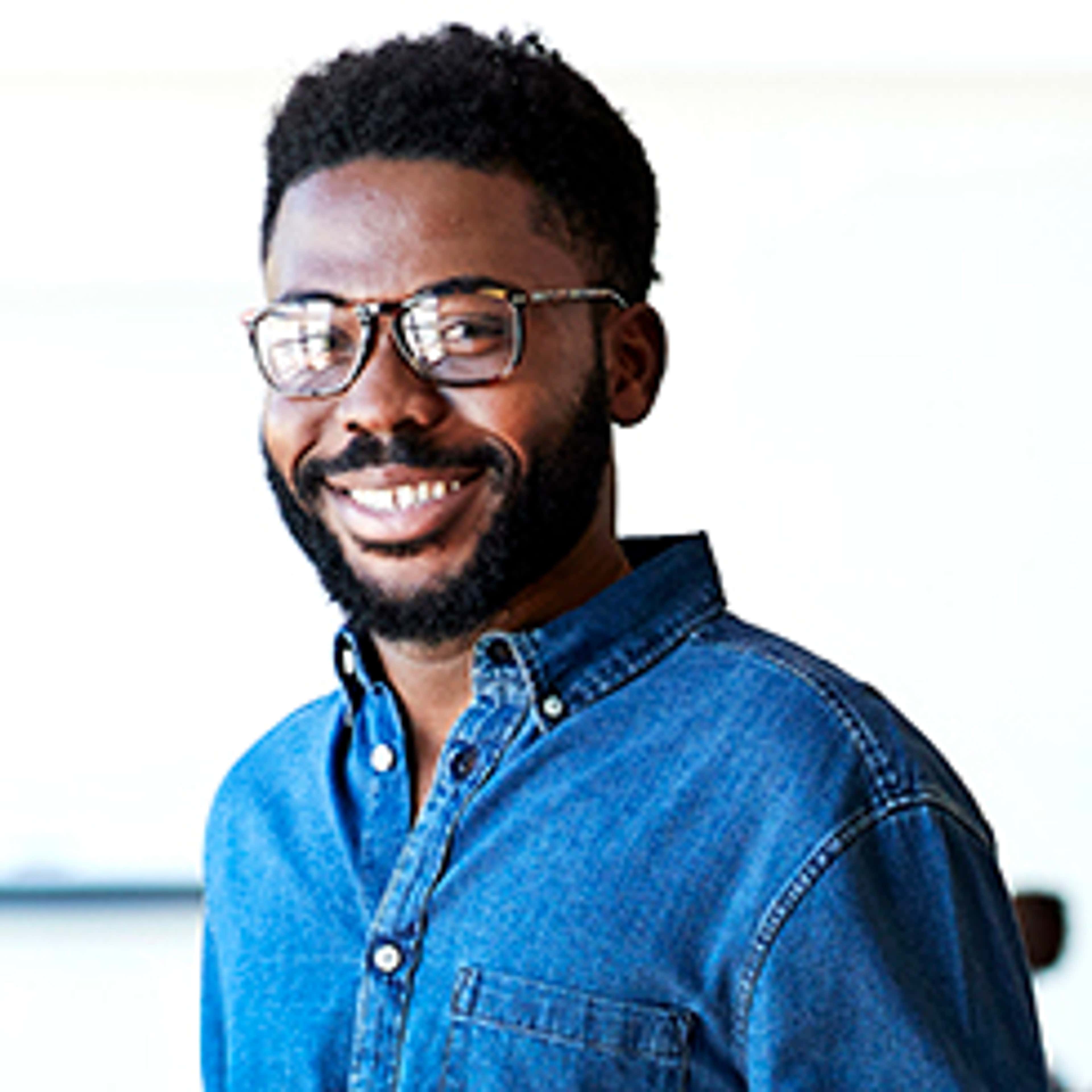 Man wearing glasses and blue denim shirt smiling into camera with white background