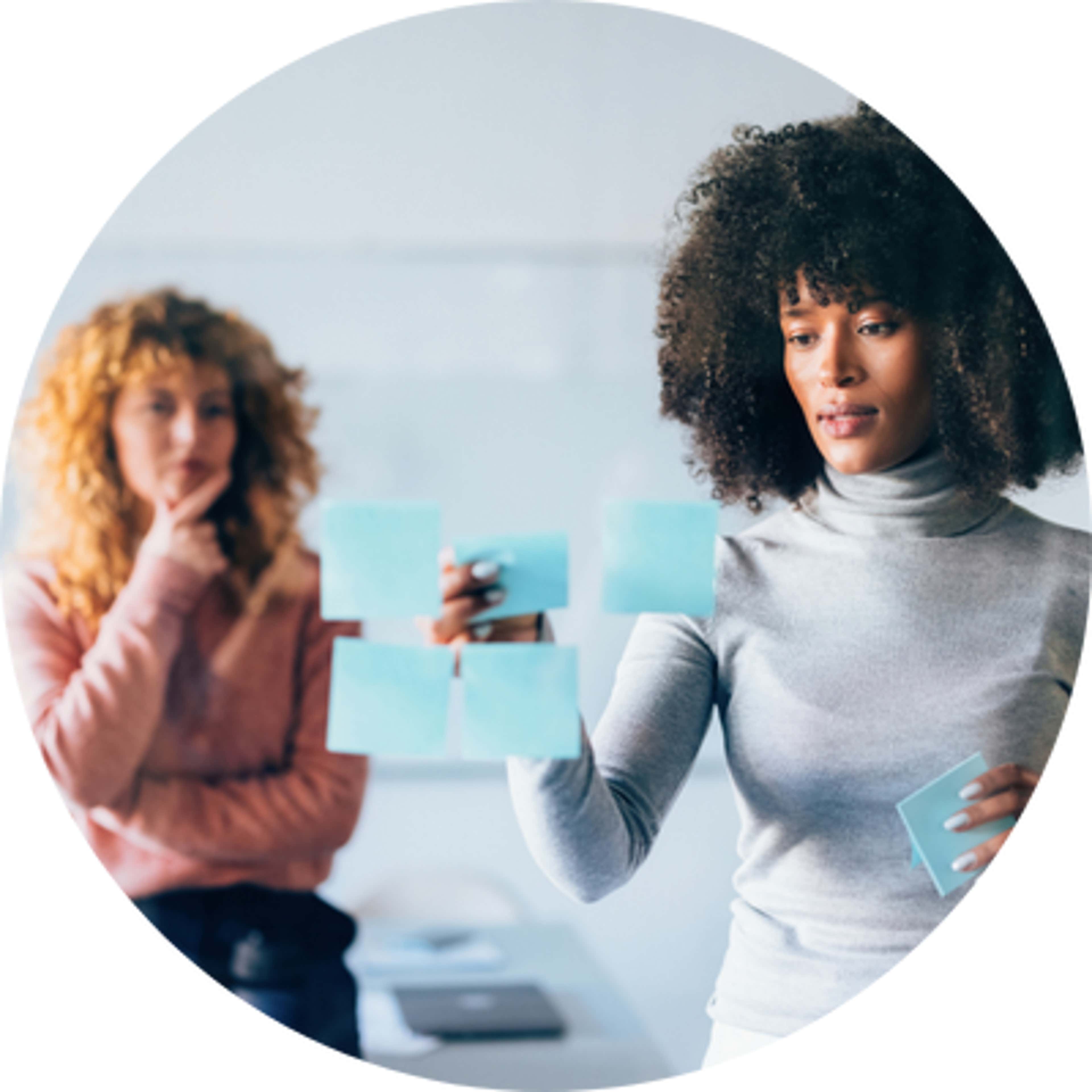 Two women standing in a room - one sticking blue sticky notes on mirror, while other observes with a hand on her chin