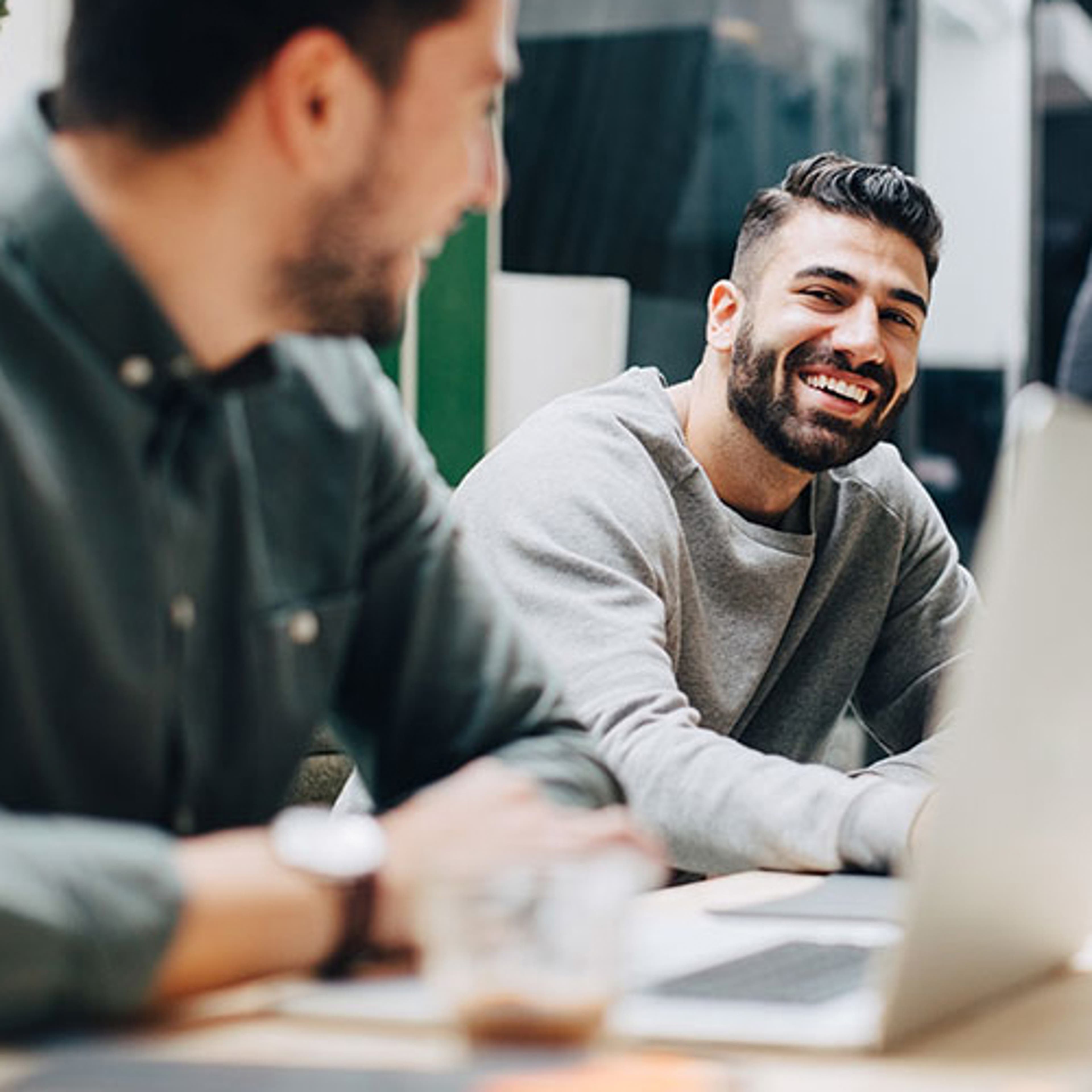 Man smiling at another man who is on his laptop