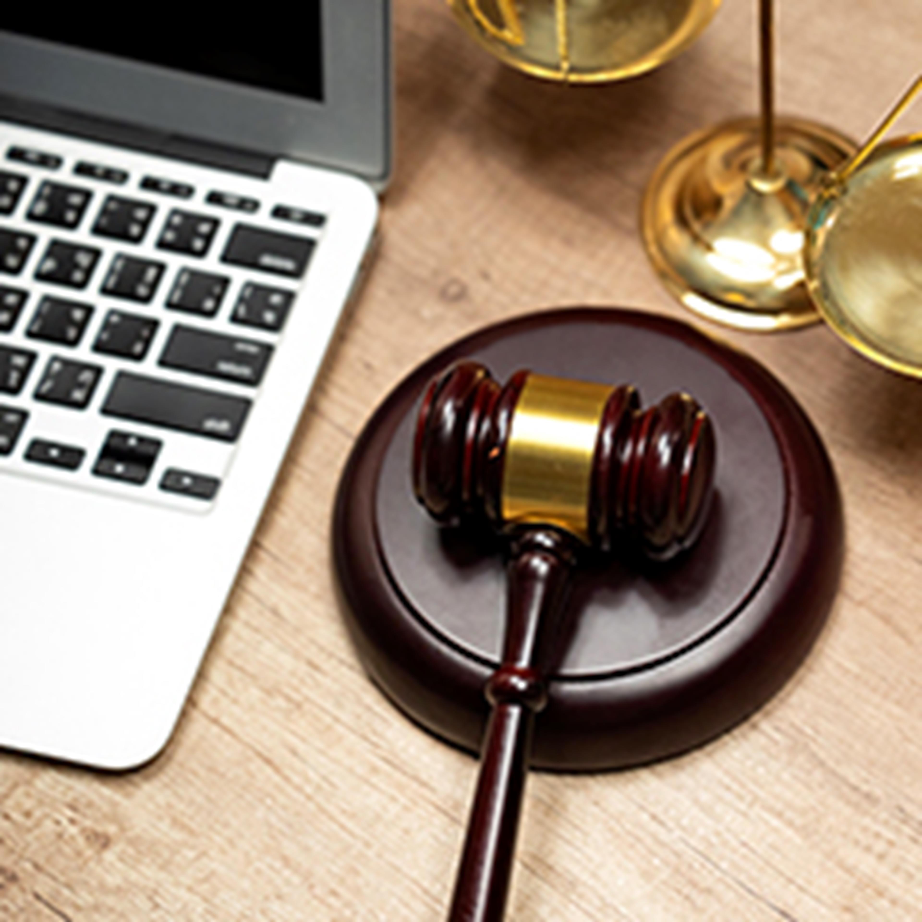 Partially visible laptop on the left of the image, a wooden gavel and block to the lower right and a partially visible set of golden scales to the top right of the image