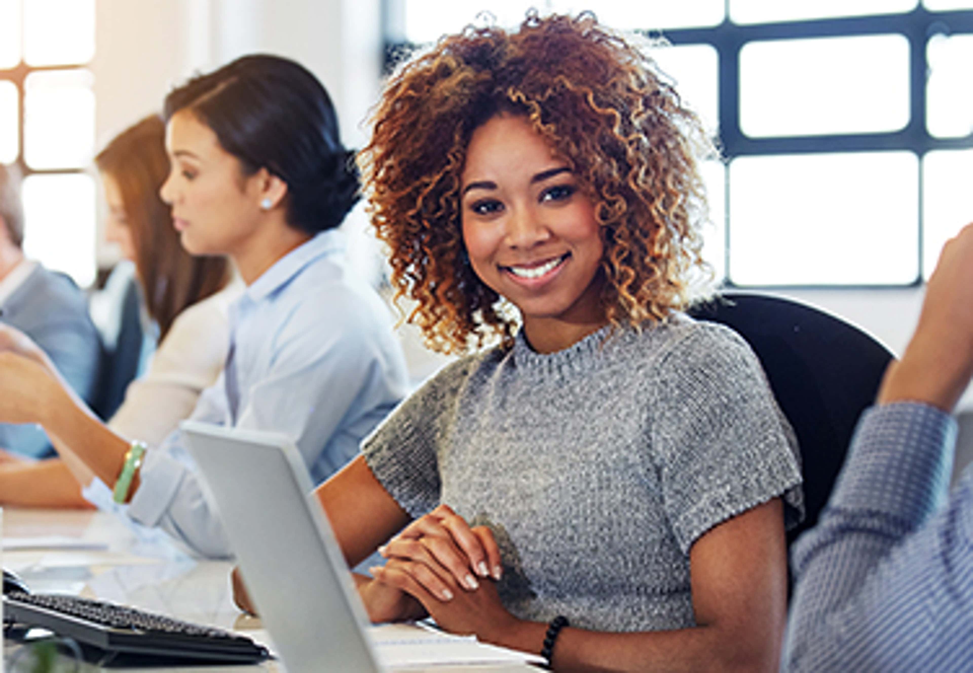 Woman smiling at camera with laptop and notes in front of her with other people in background