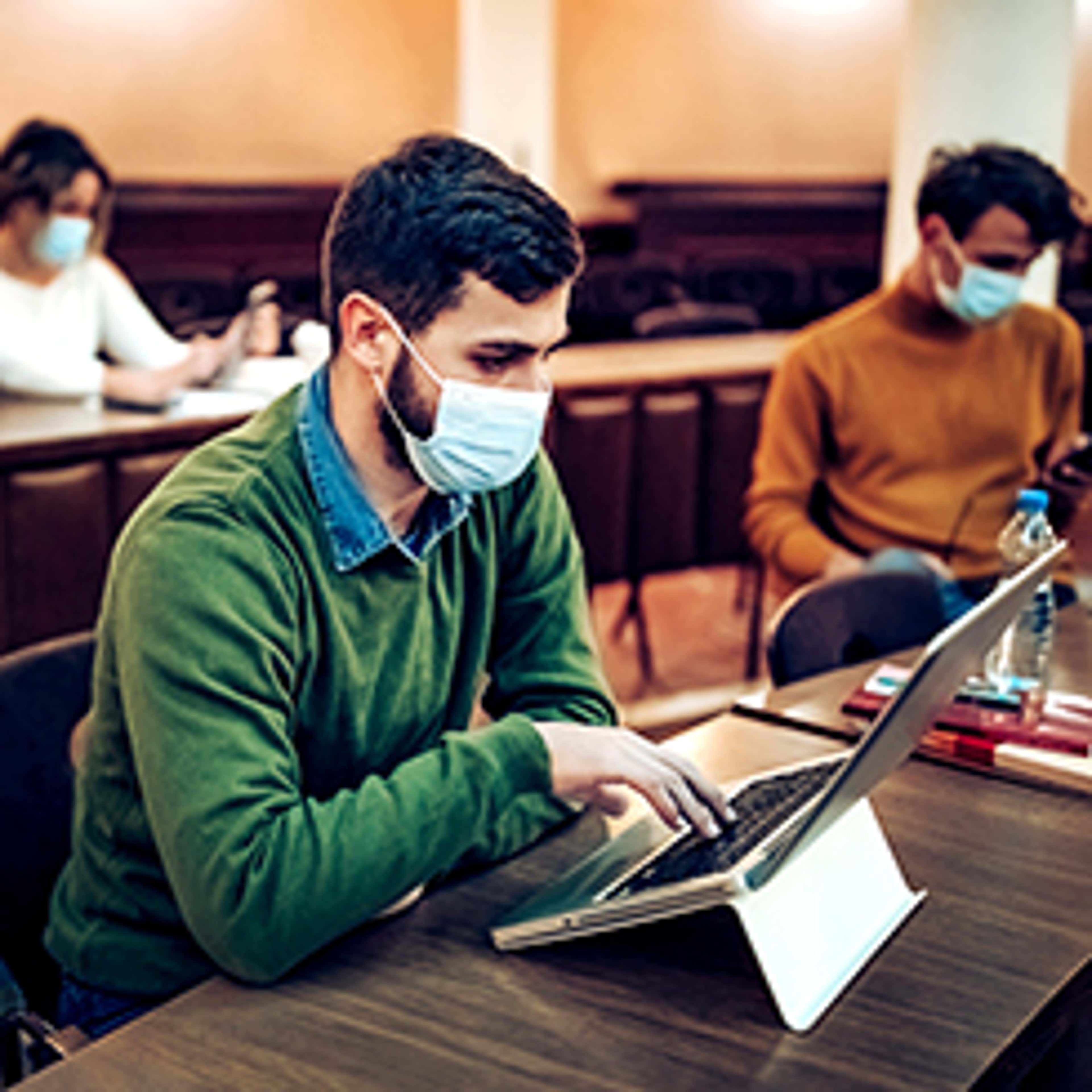Young man wearing medical mask, green sweater, working on tablet with other students in background in a study hall
