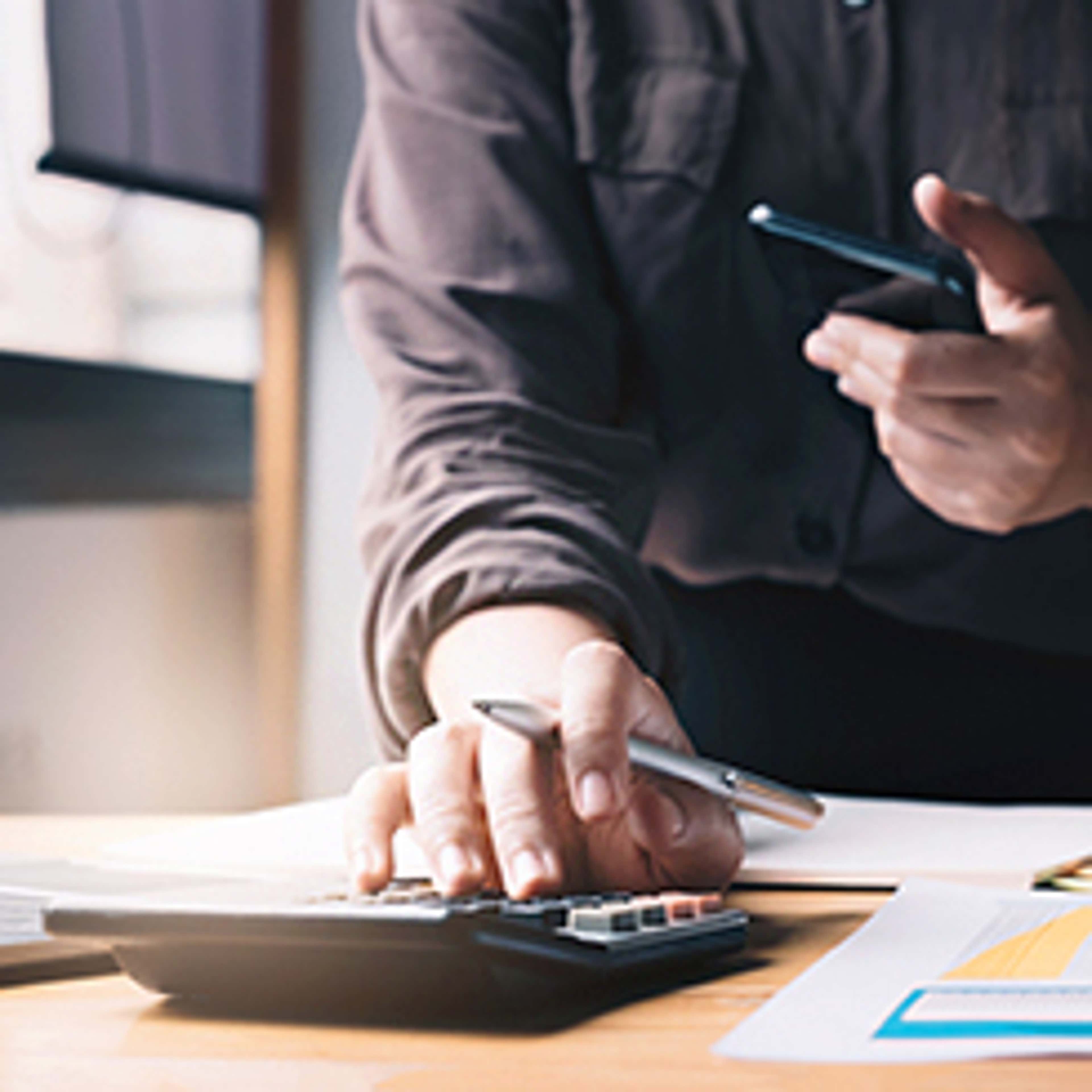 Close up of person using calculator and mobile while holding a pen while preparing for an exam
