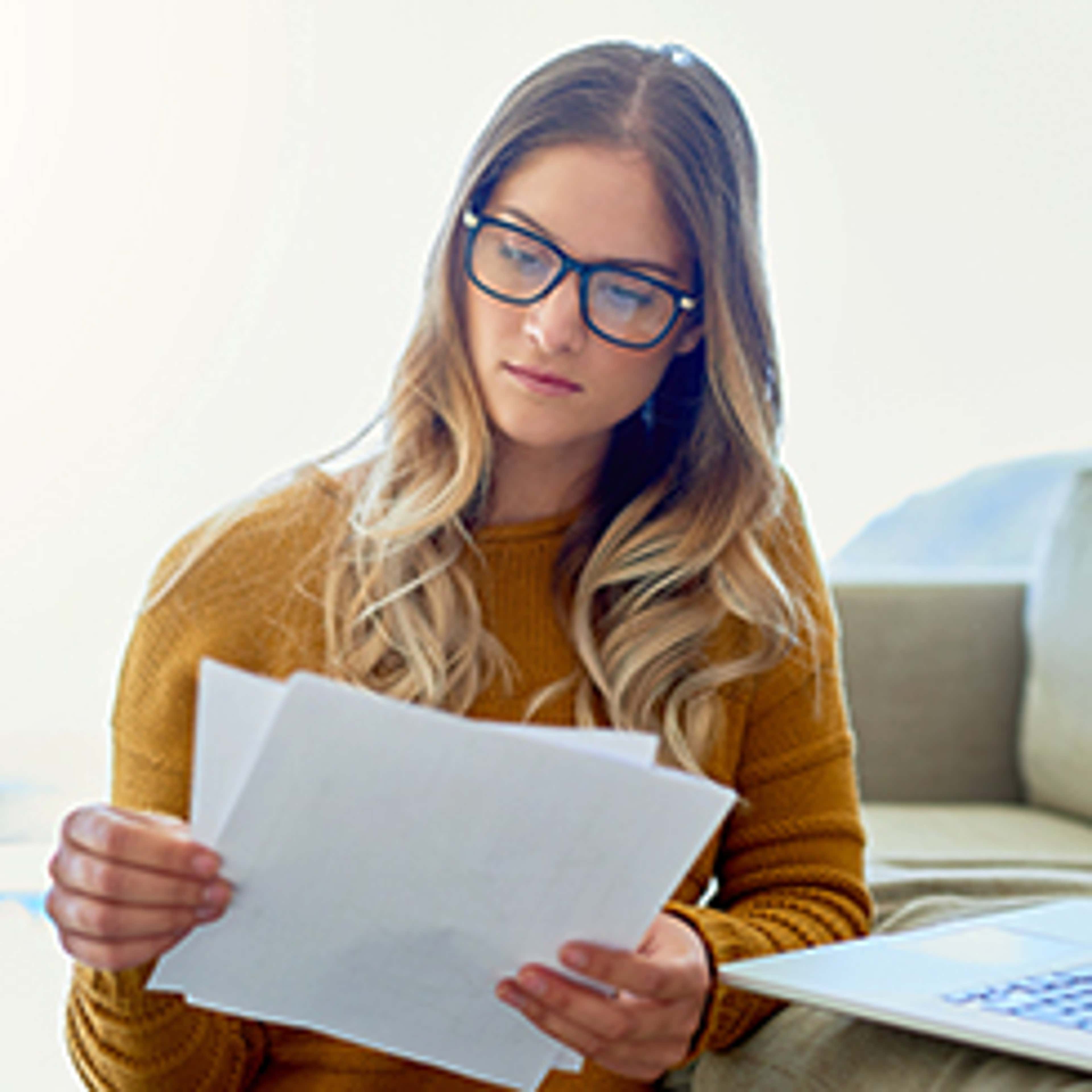 Woman wearing glasses and yellow shirt reads documents with a laptop open to the side