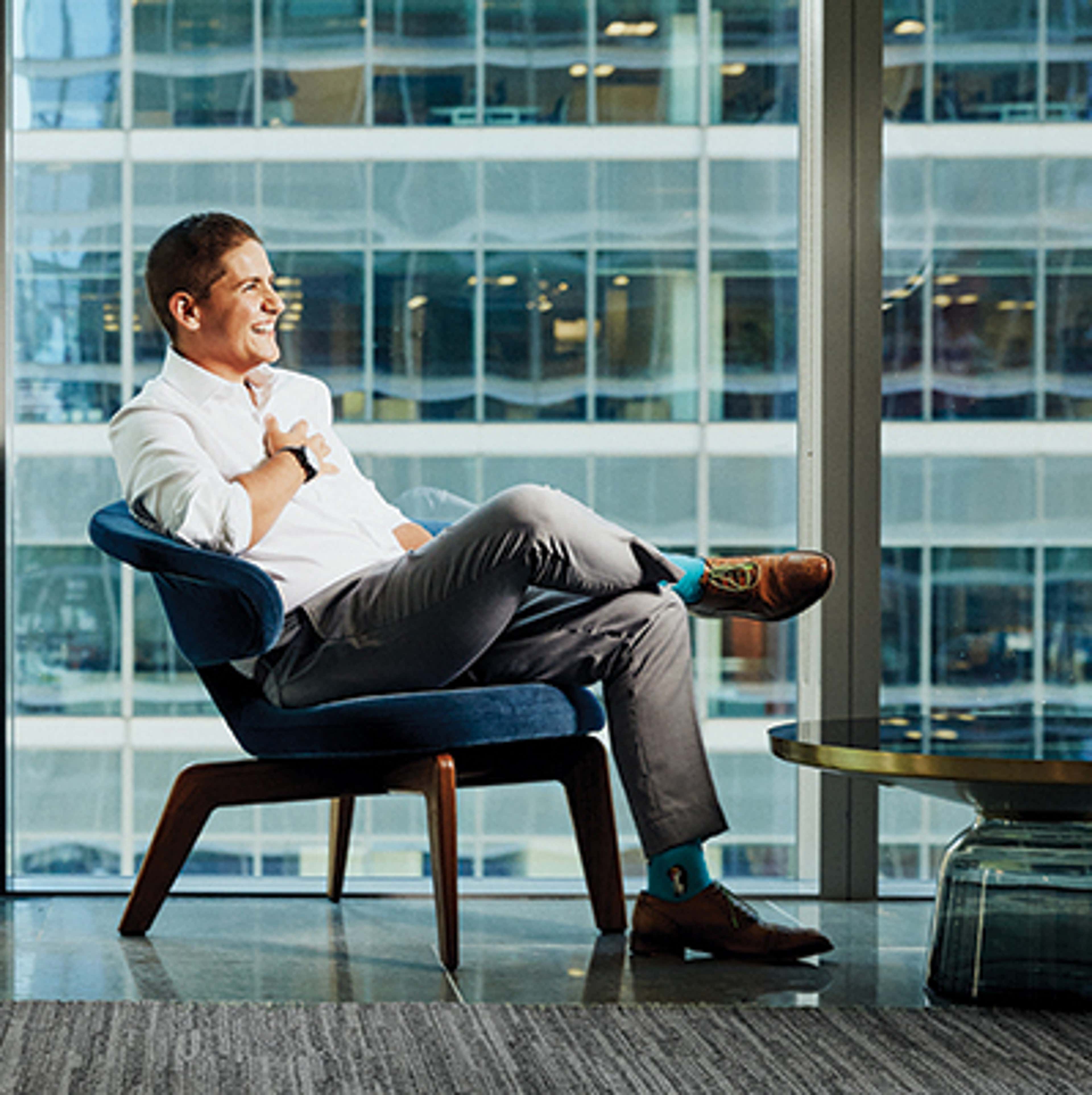 Person sitting in a chair in front of a floor to ceiling glass window, looking away from the camera and smiling